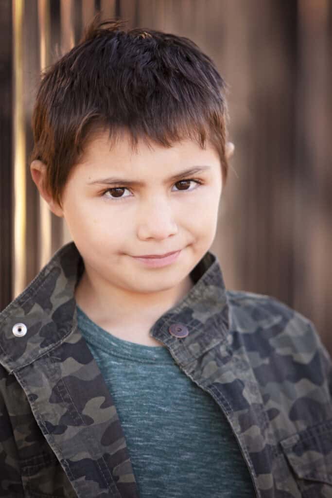 A young boy with short brown hair, wearing a green shirt and a camouflage jacket, stands outdoors in Calgary with a slight smile on his face. The softly blurred background creates a classic kids headshots vibe. - Jodi O Photography