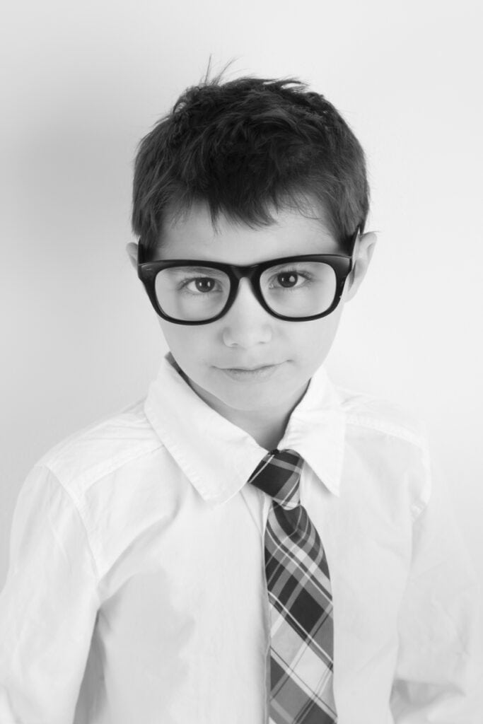 A young boy wearing large black glasses, a white button-up shirt, and a plaid tie, looking at the camera with a slight smile. This black and white portrait is perfect for Calgary kids headshots. - Jodi O Photography