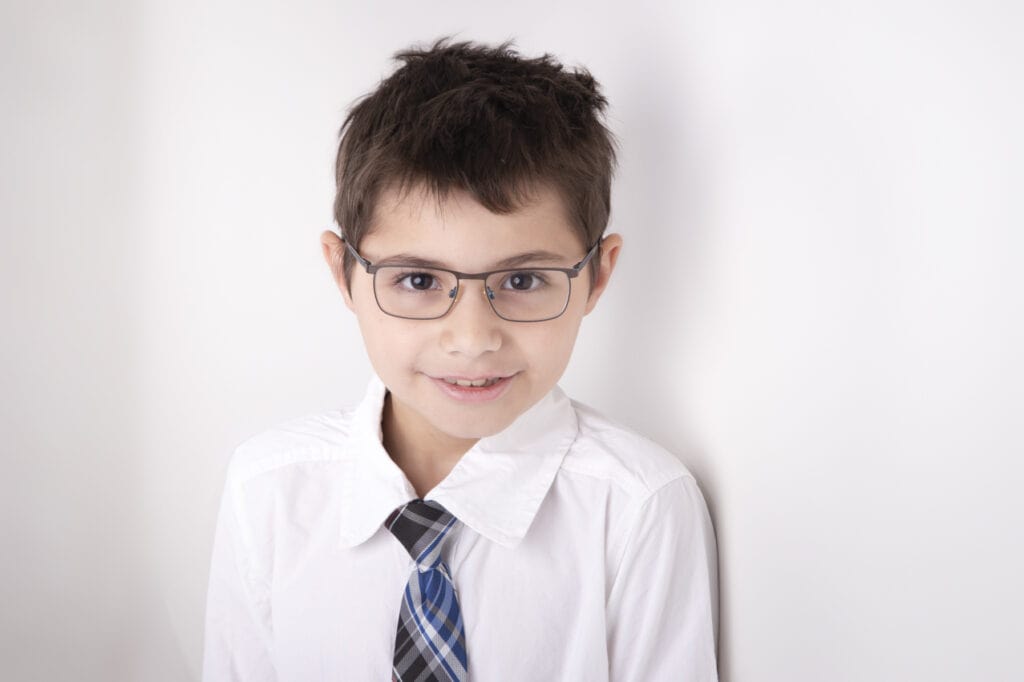 A young boy with short brown hair and glasses smiles at the camera. He is wearing a white dress shirt and a blue, black, and white striped tie, perfect for kids headshots Calgary, standing against a plain white background. - Jodi O Photography