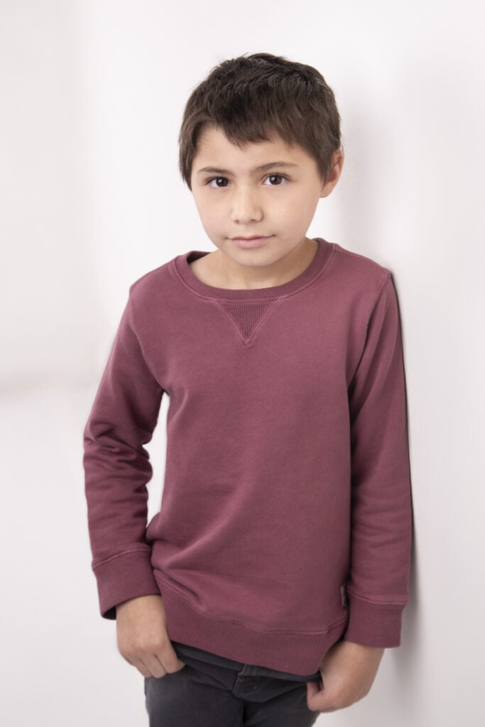 A young boy with short brown hair, wearing a mauve long-sleeve sweatshirt and black pants, stands against a white wall, looking at the camera with a soft, neutral expression—perfect for kids headshots Calgary. - Jodi O Photography