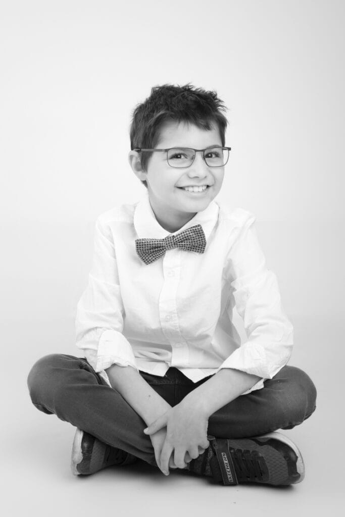 A smiling child with short hair, wearing glasses, a white shirt, a polka dot bow tie, jeans, and sneakers, sits cross-legged on the floor against a plain white background. This black and white image is perfect for kids headshots in Calgary. - Jodi O Photography