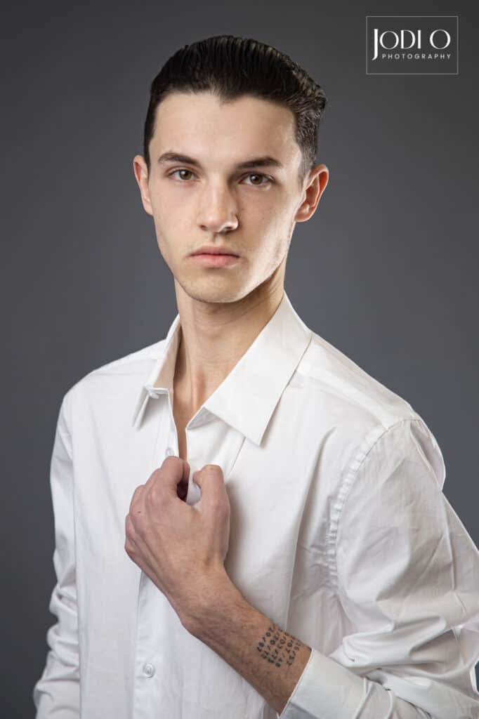 A young man with dark hair, wearing a white button-up shirt, poses against a gray background—perfect for modelling portfolios. He holds his collar to show a wrist tattoo. The image features the "Jodi O Photography" logo, known among YYC photographers. - Jodi O Photography