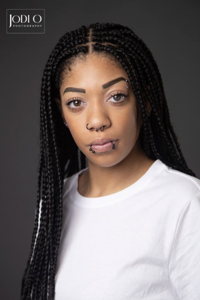 A person with long braided hair, wearing a white t-shirt, poses against a dark background—perfect for Modelling portfolios. They have multiple facial piercings and a neutral expression. The logo "Jodi O Photography" appears in the top left corner. - Jodi O Photography