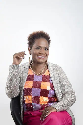 A smiling woman with short curly hair sits on a chair in bold, colorful attire. Perfect for professional business photos, her style and confidence can help enhance your brand with a modern, vibrant touch. - Jodi O Photography