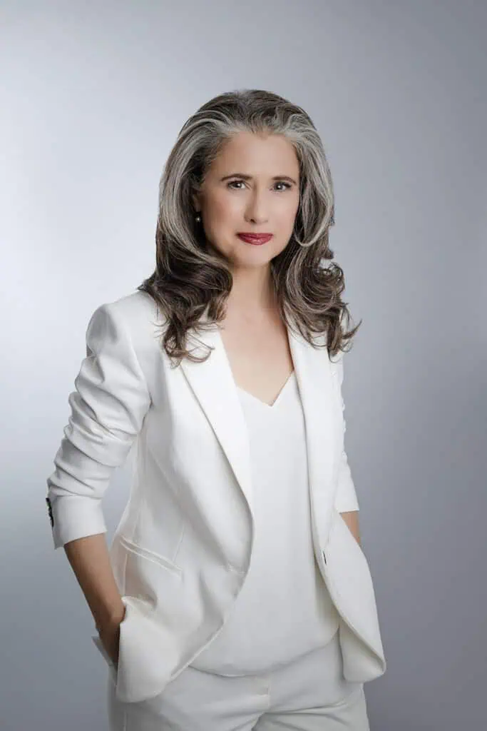 A confident woman with long, wavy gray hair wears a white suit and top, standing against a plain gray background with her hands in her pockets, looking at the camera with a slight smile—perfect for professional business photos to enhance your brand. - Jodi O Photography