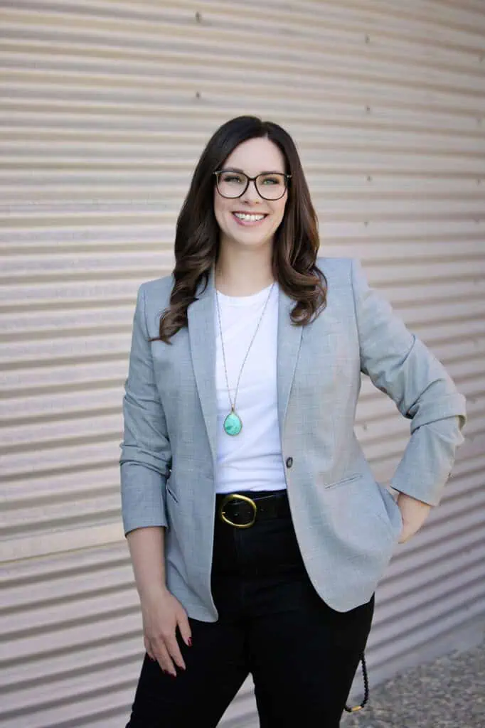 A woman with long brown hair and glasses smiles, standing in front of a corrugated metal wall. Dressed in a light gray blazer and turquoise pendant, she’s ready for professional business photos to enhance your brand. - Jodi O Photography