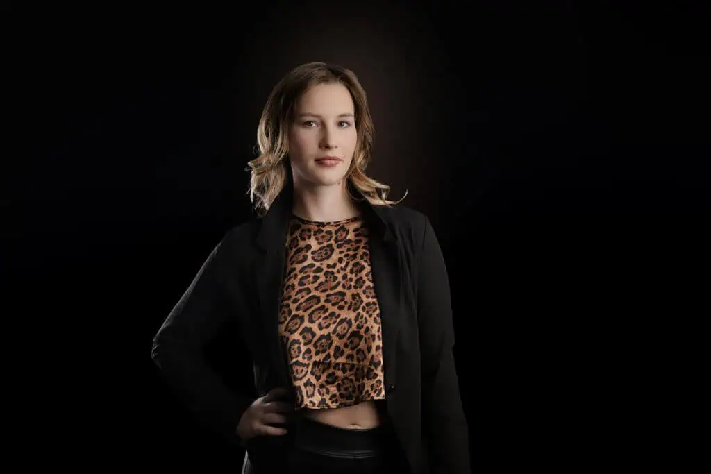 A woman with wavy blonde hair poses confidently with one hand on her hip, wearing a black blazer over a leopard print top, against a plain black background—perfect for professional business photos to enhance your brand. - Jodi O Photography
