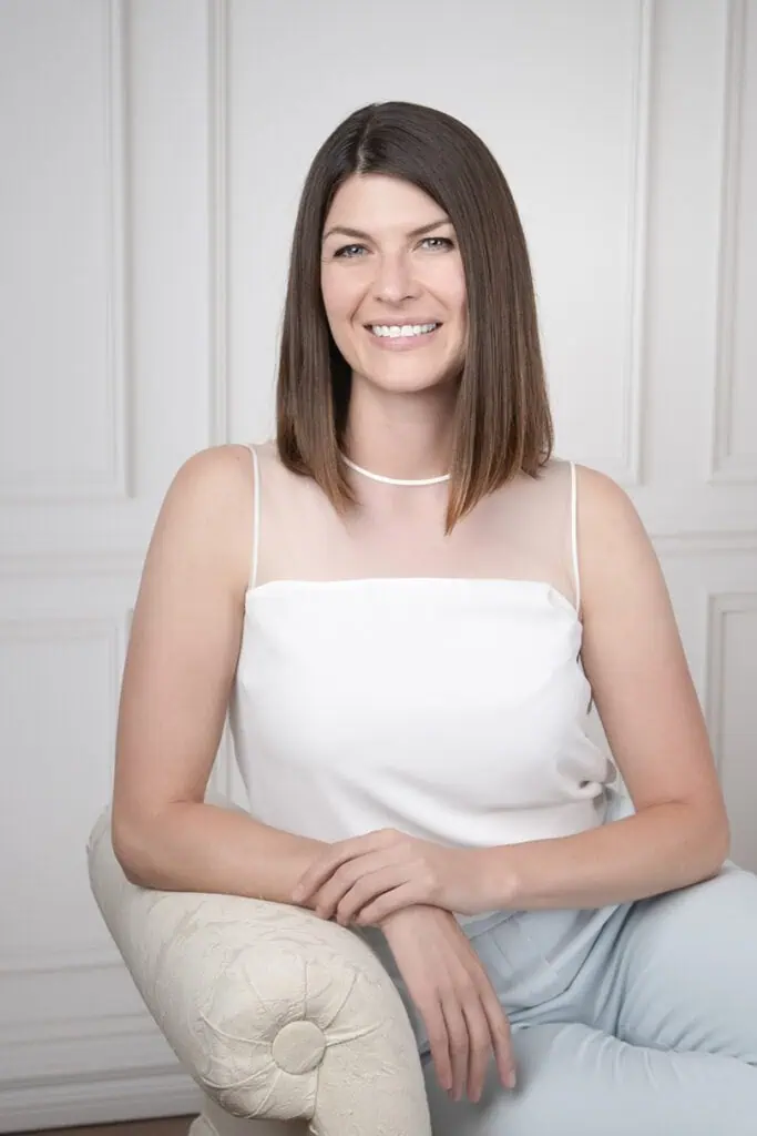 A woman with straight brown hair, wearing a sleeveless white top and light blue pants, sits on a cream-colored chair and smiles at the camera in a bright, elegantly styled room—perfect for professional business photos to enhance your brand. - Jodi O Photography