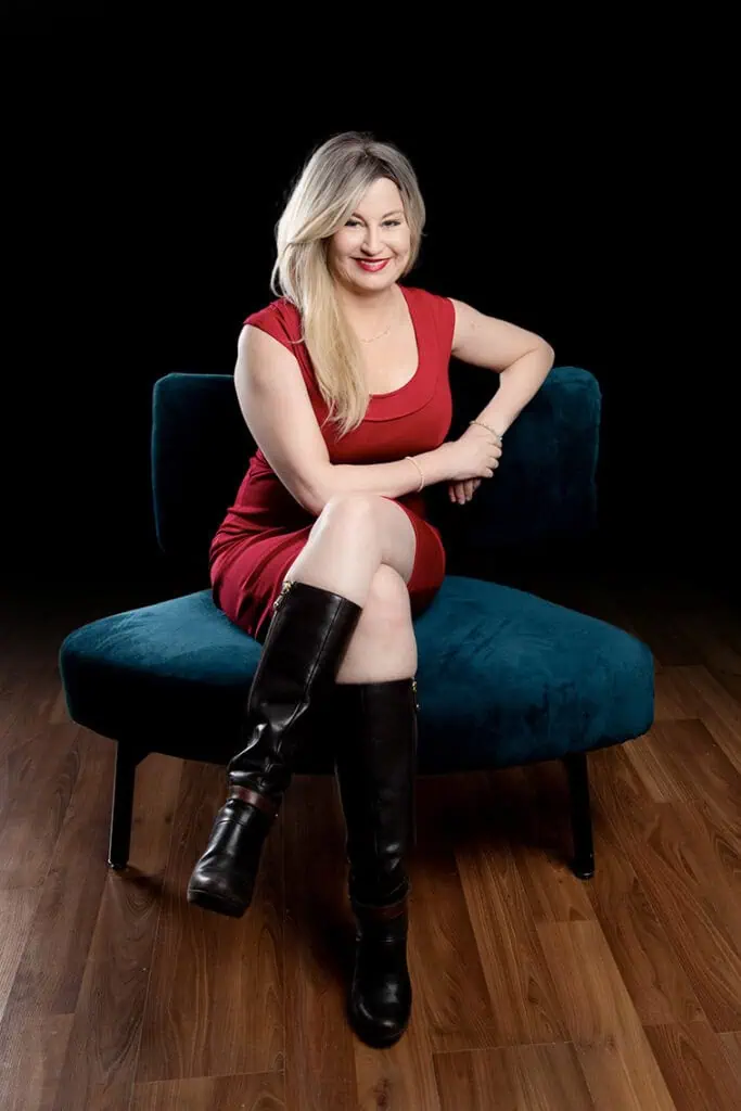 A woman with long blonde hair in a red dress and black knee-high boots sits cross-legged on a blue velvet chair against a dark background, smiling confidently—perfect for professional business photos to enhance your brand. - Jodi O Photography