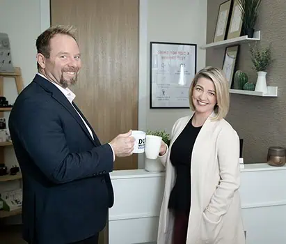 Two professionally dressed people stand in an office, smiling and holding coffee mugs. Shelves with decor, framed certificates, and a wooden door are visible in the background—perfect for professional business photos to enhance your brand. - Jodi O Photography