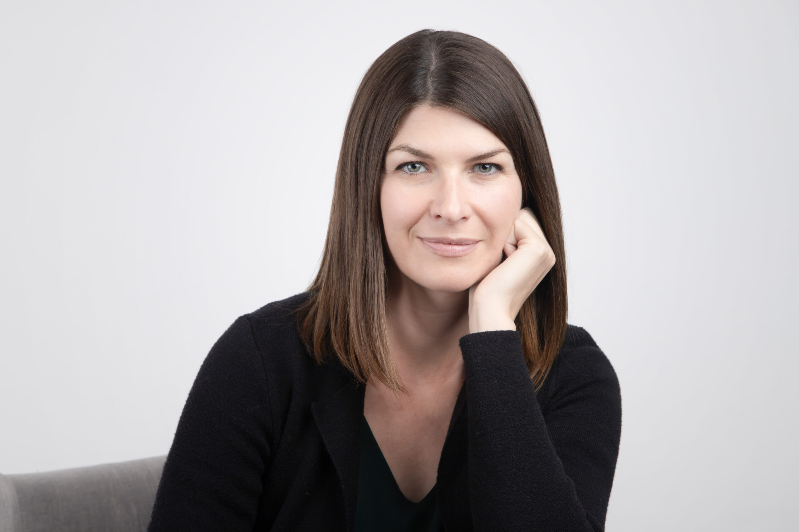 A woman with straight brown hair and fair skin sits on a chair, resting her head on her hand, and smiles gently at the camera. She is wearing a black sweater against a plain light background, perfect for showcasing Calgary headshots. - Jodi O Photography