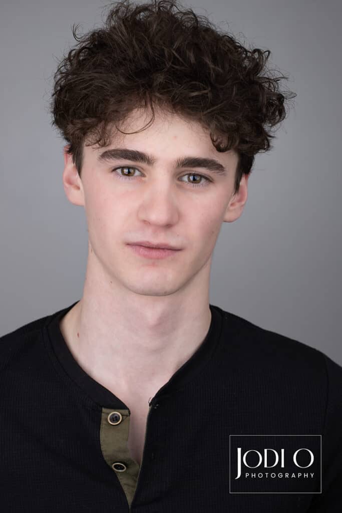 A young man with curly dark hair and a slight smile faces the camera against a plain gray background, making this image ideal for modelling portfolios. He is wearing a black shirt. The logo "Jodi O Photography" is visible in the bottom right corner. - Jodi O Photography