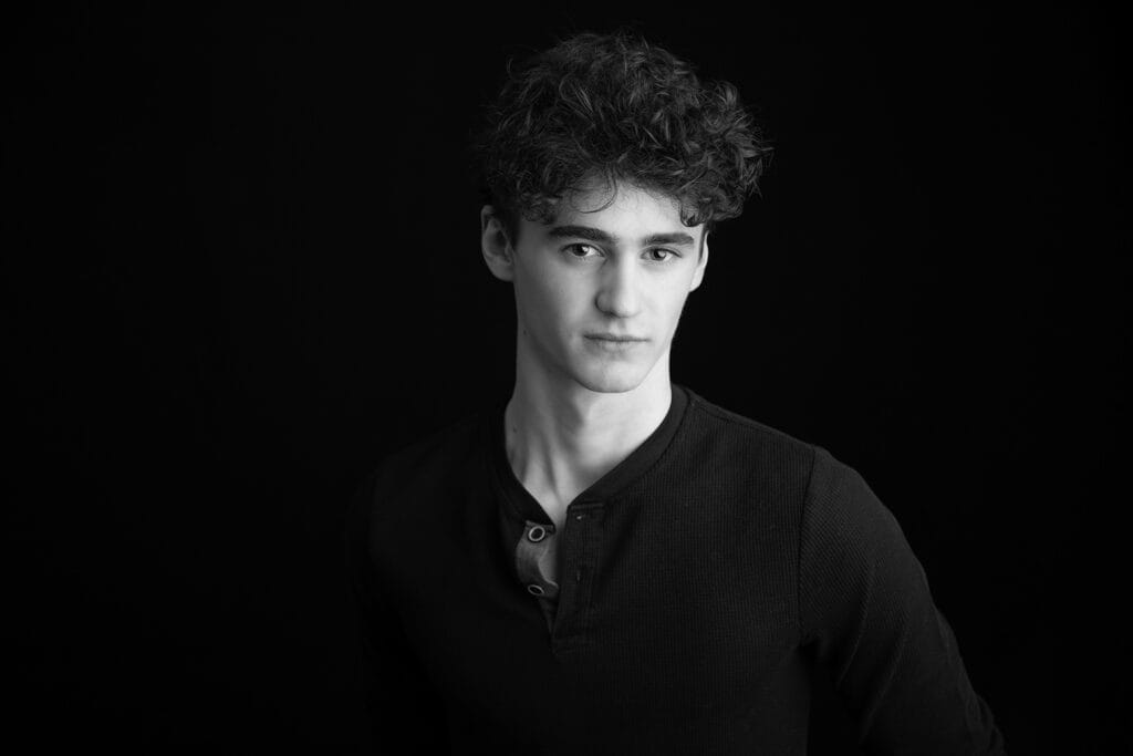 A young man with curly hair wearing a dark, long-sleeved shirt stands against a plain black background, looking slightly to the side with a neutral expression—an ideal shot for modelling portfolios or YYC photographers seeking striking portraits. - Jodi O Photography
