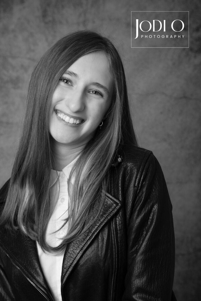 A young woman with long straight hair smiles at the camera, wearing a leather jacket over a buttoned shirt. Perfect for Modelling Portfolios, this black and white photo by YYC Photographers features a textured backdrop and “Jodi O Photography” logo. - Jodi O Photography