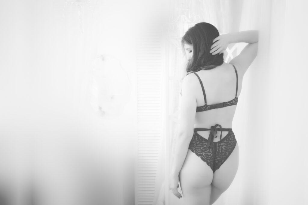 A woman wearing sexy black lingerie stands with her back to the camera in a softly lit, white studio, looking over her shoulder with one hand in her hair. The dreamy black and white image captures an ethereal moment to celebrate confidence. - Jodi O Photography