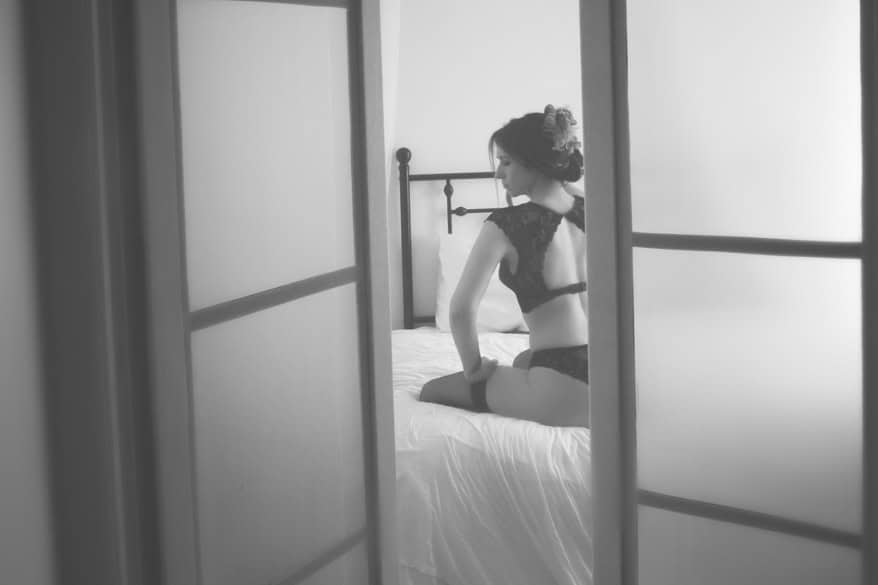 A woman in lace lingerie sits on a bed in a softly lit room, viewed through partially open sliding doors. This black and white studio photography captures a gentle, intimate atmosphere—a perfect way to celebrate yourself. - Jodi O Photography