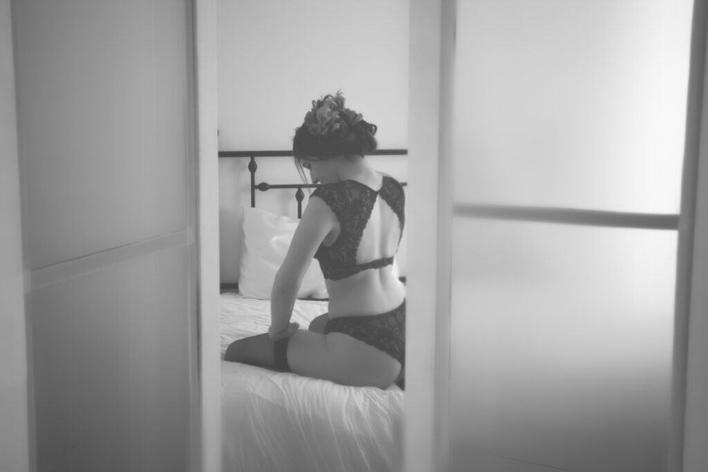 A woman in black lace lingerie and stockings sits on a bed, facing away, with soft light coming through frosted glass panels. This black and white studio photography captures a moment to celebrate your sexy side. - Jodi O Photography
