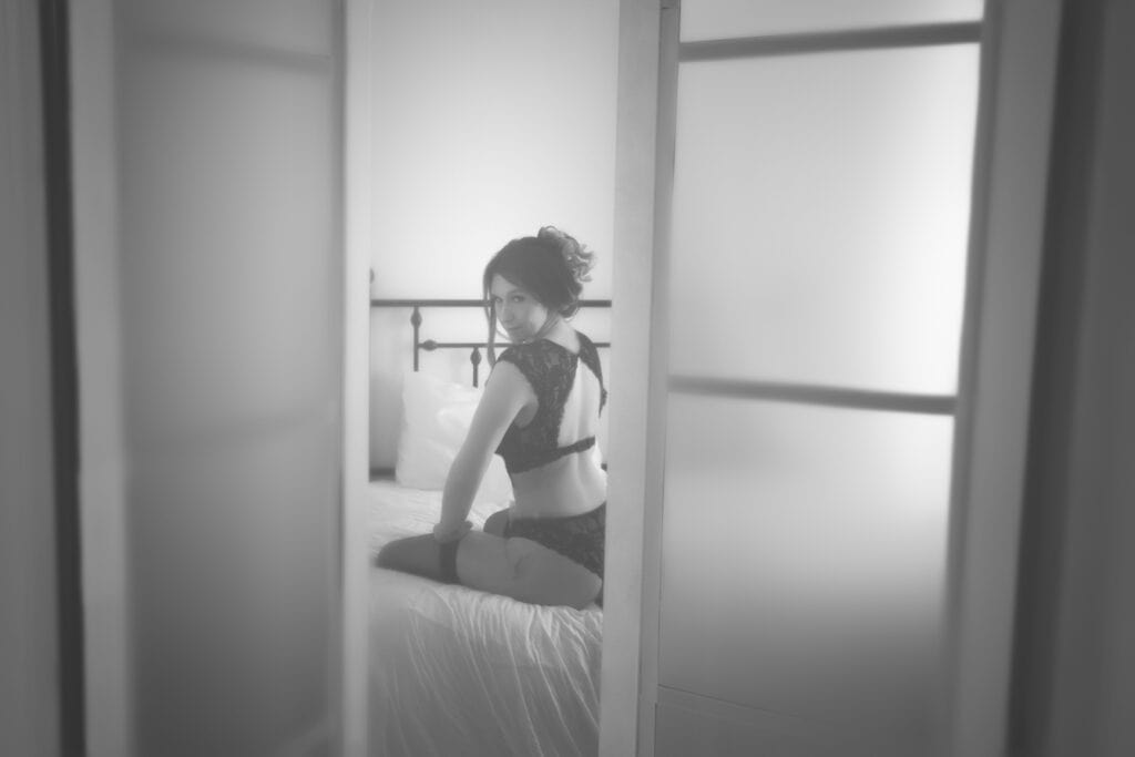 A woman in sexy black lingerie sits on a bed, looking over her shoulder at the camera. The scene is viewed through slightly open sliding doors, with soft, diffused lighting creating a gentle, intimate atmosphere. - Jodi O Photography