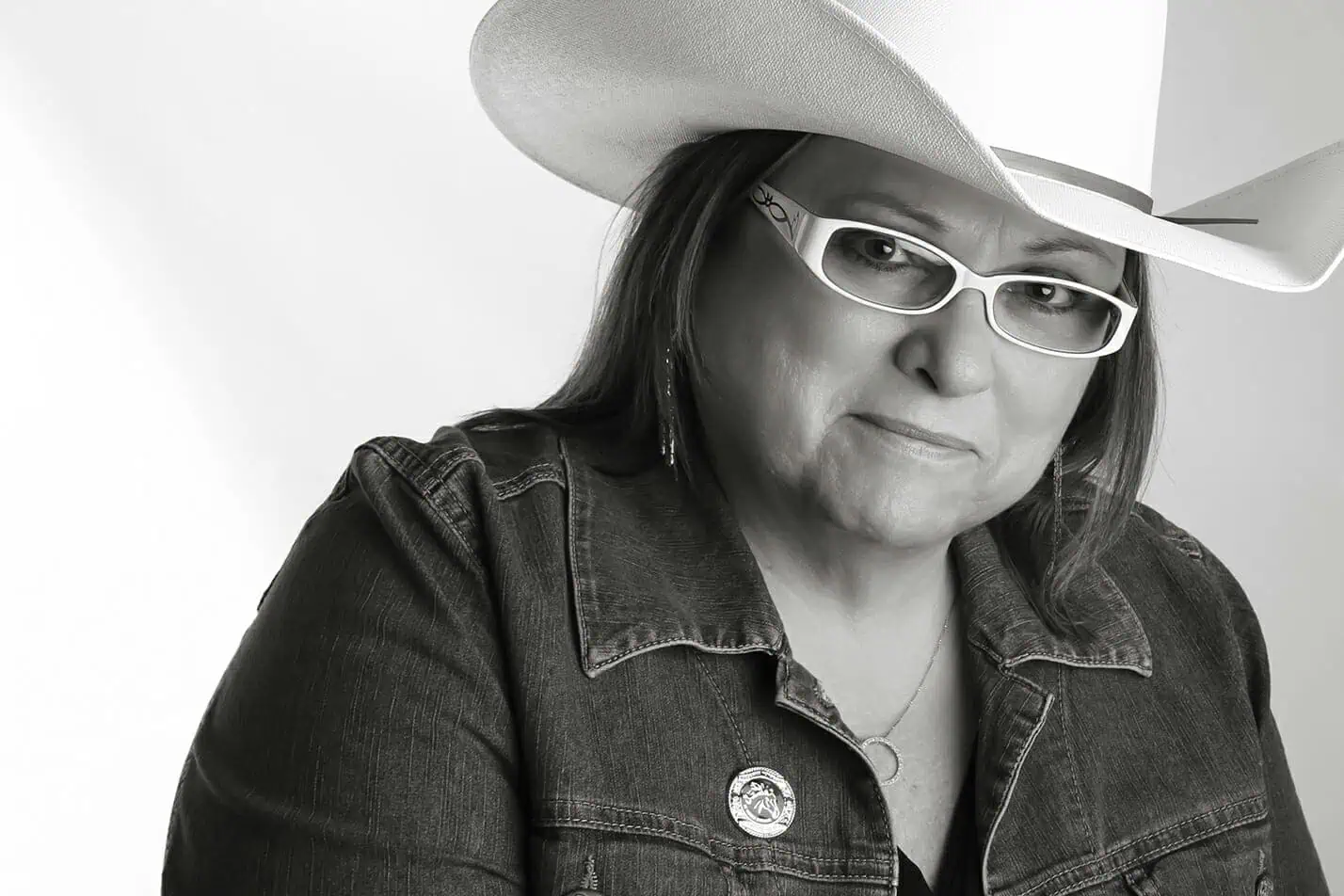 Black and white photo of a person in a cowboy hat, white glasses, denim jacket with a pin, and necklace, offering a slight smile—perfect for those seeking unique professional headshots Calgary style. - Jodi O Photography
