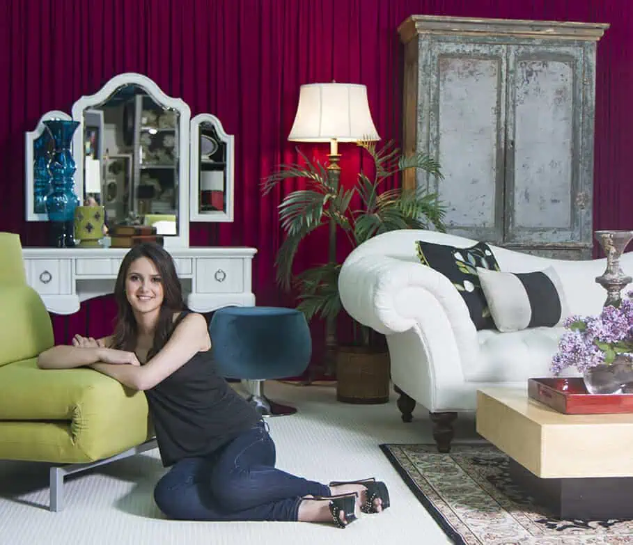 A woman with long dark hair sits on the floor, leaning against a green sofa in a stylish living room—perfectly staged like those seen in professional headshots Calgary photographers capture, with a white couch, antique armoire, vanity mirror, plants, and red curtains. - Jodi O Photography