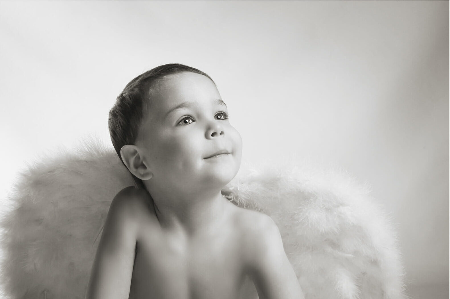 A young child with short hair smiles and looks upward, sitting in front of large, fluffy angel wings. Captured in black and white with a soft, light background—perfect for family photography Calgary moments. - Jodi O Photography