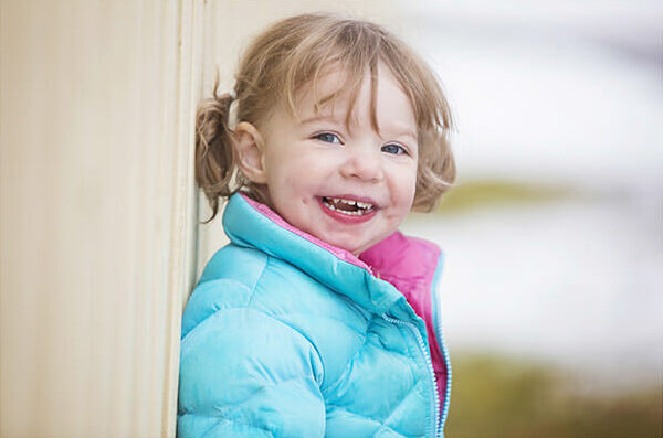Smiling young child with light brown hair in pigtails, wearing a turquoise puffy jacket with a pink lining, stands outdoors by a light-colored wall. Perfect for family photography Calgary, the softly blurred background highlights her joyful expression. - Jodi O Photography
