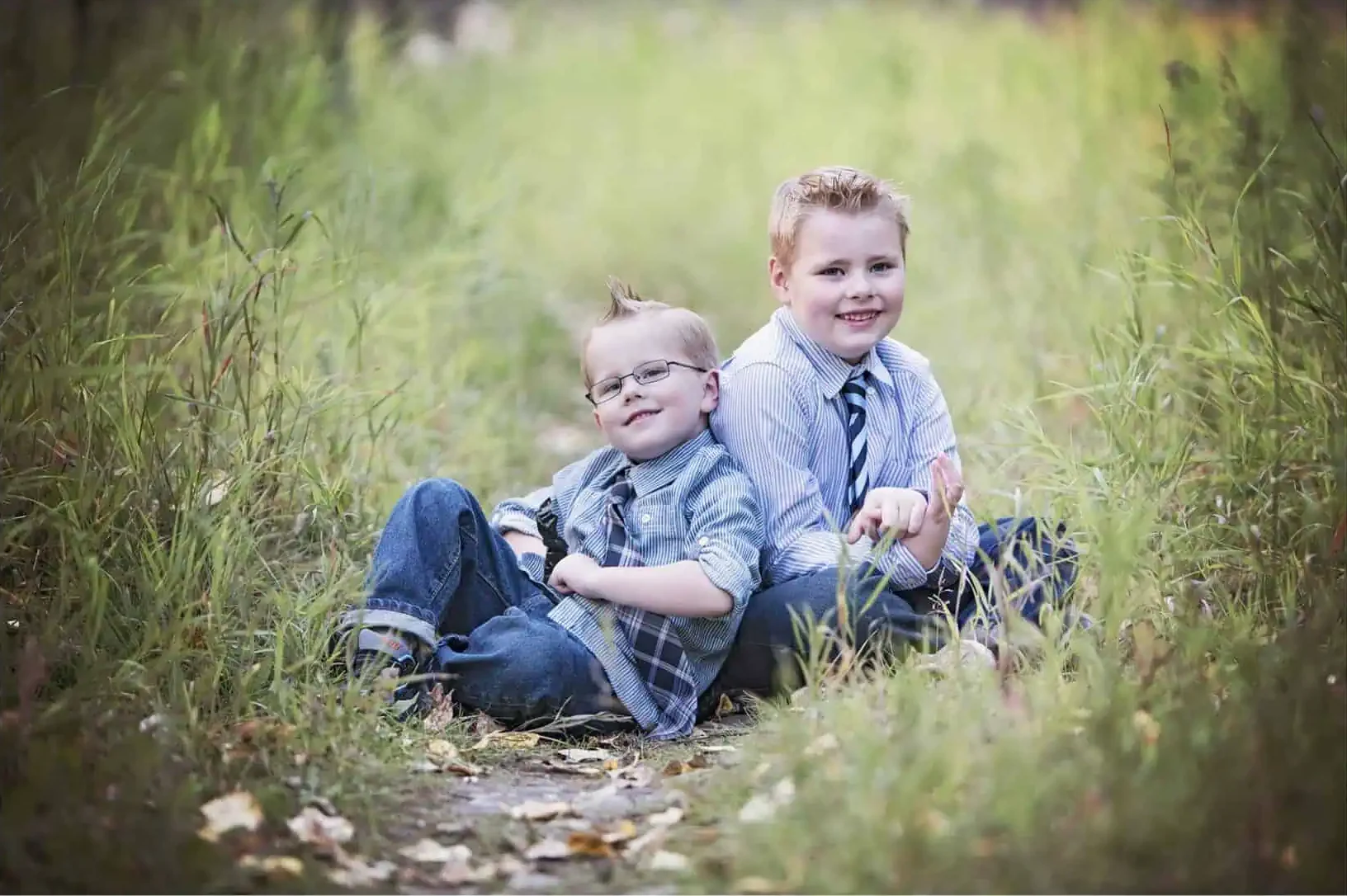 Two young boys sit back-to-back on a grassy path, smiling at the camera. Perfect for family photography Calgary, both wear jeans and collared shirts—one with glasses and plaid, the other in a tie—framed by vibrant green grass and leaves. - Jodi O Photography