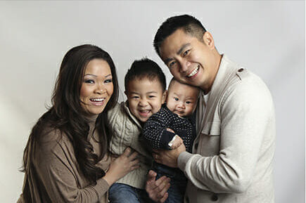 A smiling family of four poses together against a plain background. Two adults and two young children, one held by the father and the other sitting between the parents, all appear happy and close—perfect for family photography Calgary moments. - Jodi O Photography
