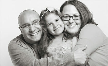 A man, a young girl, and a woman smile and hug closely together, posing for a timeless black and white family photography Calgary portrait against a plain background. - Jodi O Photography