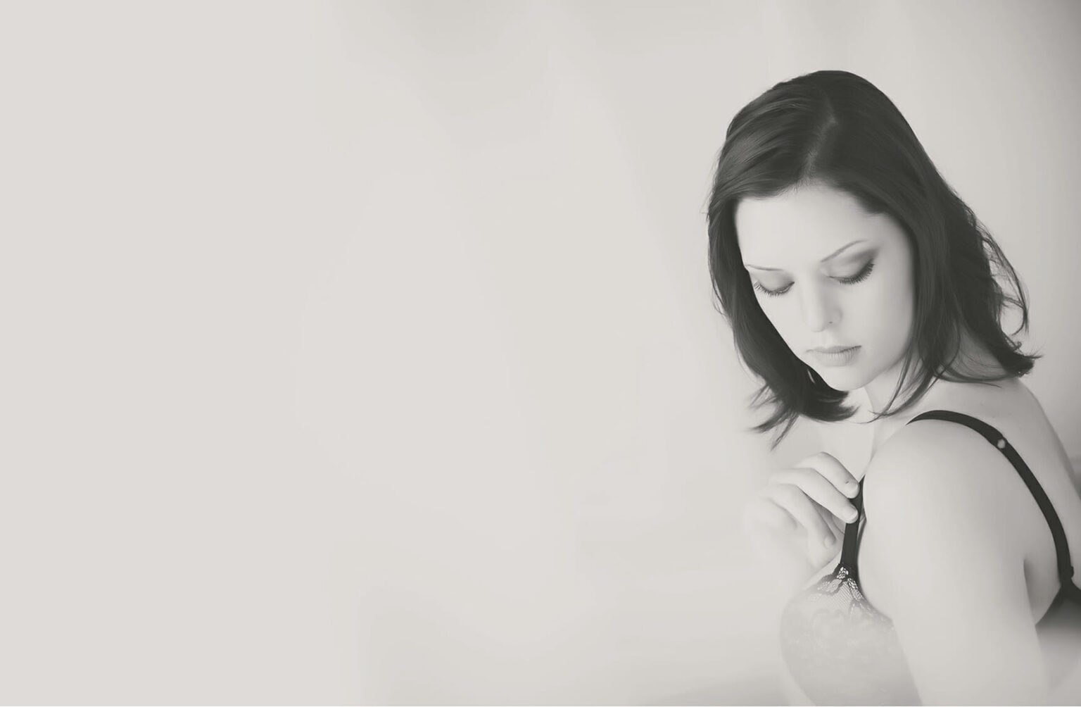 A woman with shoulder-length dark hair looks down while adjusting her bra strap, set against a soft, light background in a black-and-white photo by a boudoir photographer Calgary. - Jodi O Photography