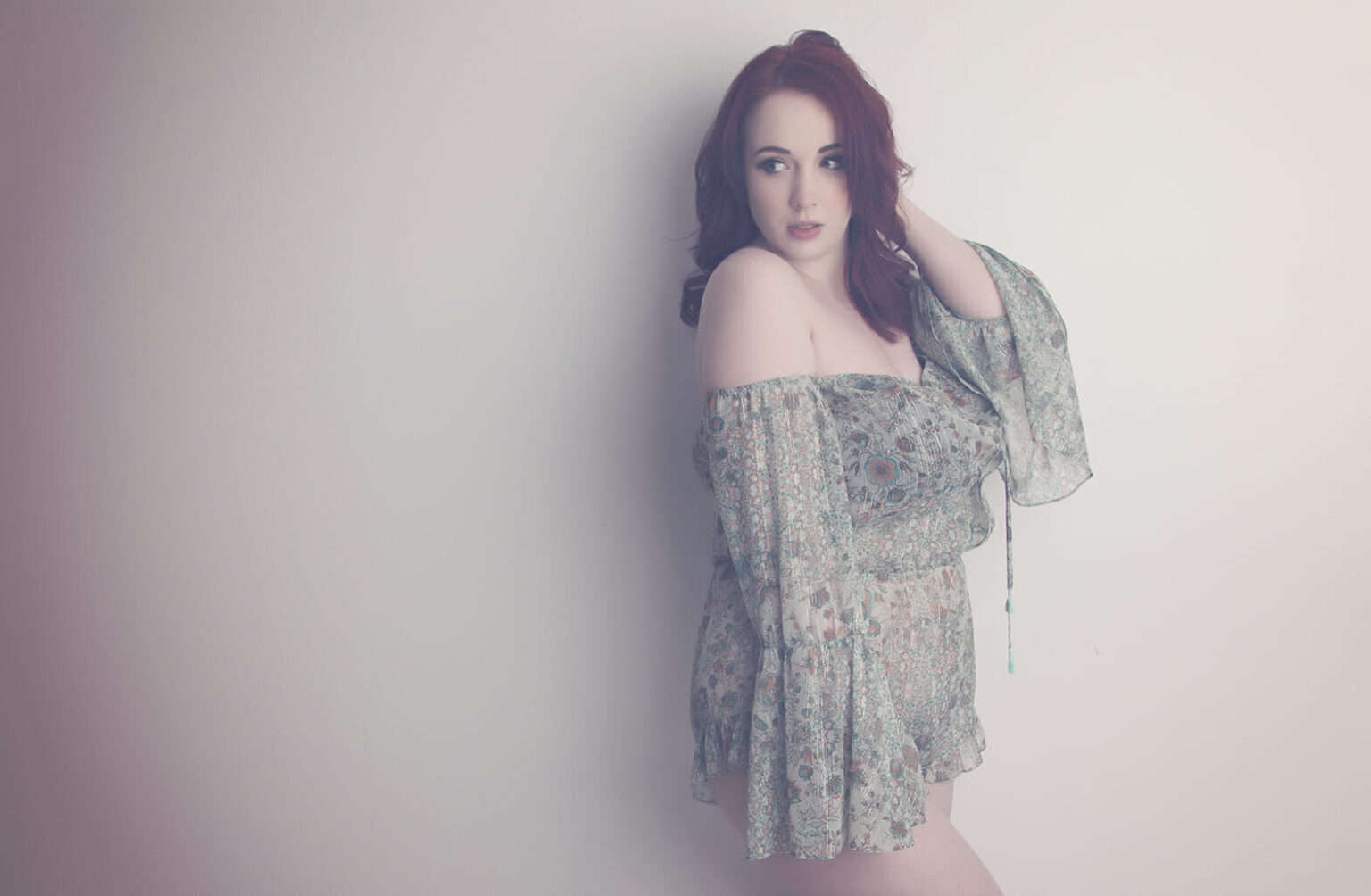 A woman with long red hair poses against a pale wall, wearing an off-the-shoulder, patterned romper with flowing sleeves. Captured by a boudoir photographer Calgary, the image features soft pastel tones and a gentle, dreamy vibe. - Jodi O Photography