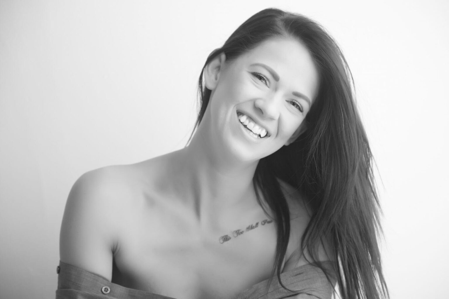 A young woman with long dark hair smiles warmly at the camera. She wears an off-the-shoulder top, revealing a small collarbone tattoo. Captured by a boudoir photographer Calgary, the black and white image features a soft, bright background. - Jodi O Photography