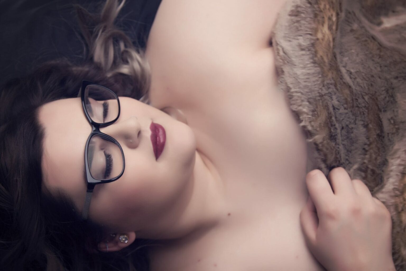 A woman with long brown hair and glasses lies on her back, eyes closed, partially covered by a soft brown blanket. Captured by a boudoir photographer in Calgary, she holds the blanket with one hand, wearing dark lipstick and a calm, relaxed expression. - Jodi O Photography