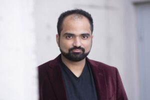 A man with a trimmed beard and short hair, wearing a maroon blazer over a black shirt, stands in front of a light gray background, looking directly at the camera with a neutral expression—perfect for actor headshots Calgary. - Jodi O Photography