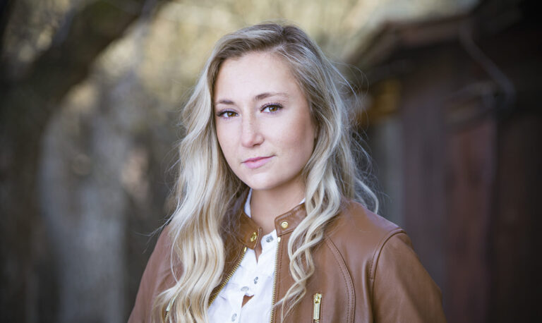 A woman with long blonde hair wearing a brown leather jacket and white shirt stands outdoors, looking confidently at the camera with a slight smile—a style ideal for actor headshots Calgary professionals seek. Blurred trees and a wooden structure form the backdrop. - Jodi O Photography
