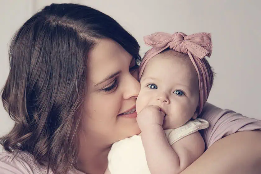 A woman with brown hair smiles while holding a baby with blue eyes and a pink headband. The baby has a hand in their mouth and both are facing the camera, capturing a sweet moment perfect for family photography Calgary style. - Jodi O Photography