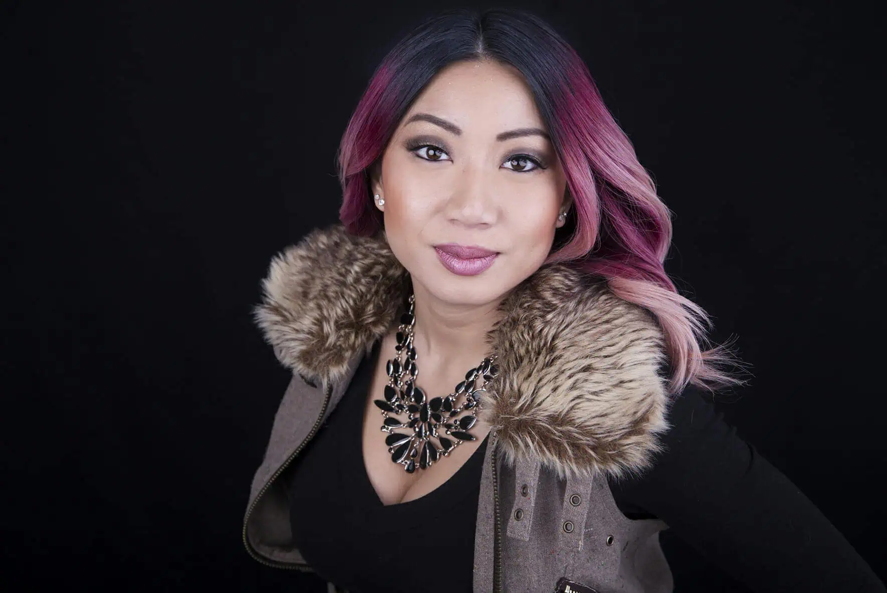 Woman with ombre pink hair and makeup poses against a black background, wearing a fur-collared vest, black top, and a large statement necklace—an inspiring look for professional headshots Calgary clients will love. - Jodi O Photography