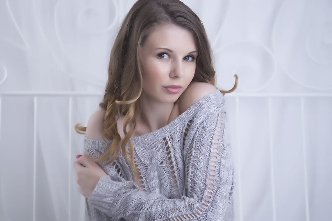 A young woman with long, wavy brown hair and light makeup wears a loose gray knit sweater off one shoulder, sitting against a softly blurred white background, gazing at the camera in a style often captured by a boudoir photographer Calgary. - Jodi O Photography
