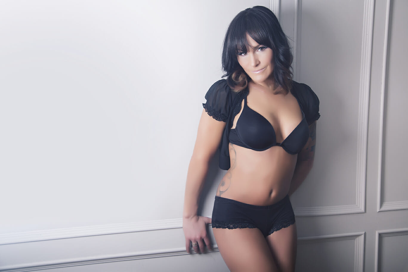 A woman with dark hair and bangs poses against a white wall, wearing a black bra and lace-trimmed shorts with a cropped black top—an elegant look often captured by a talented boudoir photographer Calgary clients adore. - Jodi O Photography