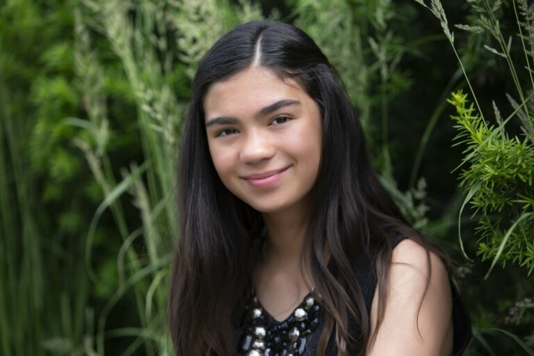 A young person with long dark hair and a sleeveless black top smiles softly while sitting outdoors among tall green plants, capturing the natural vibe perfect for actor headshots Calgary. - Jodi O Photography