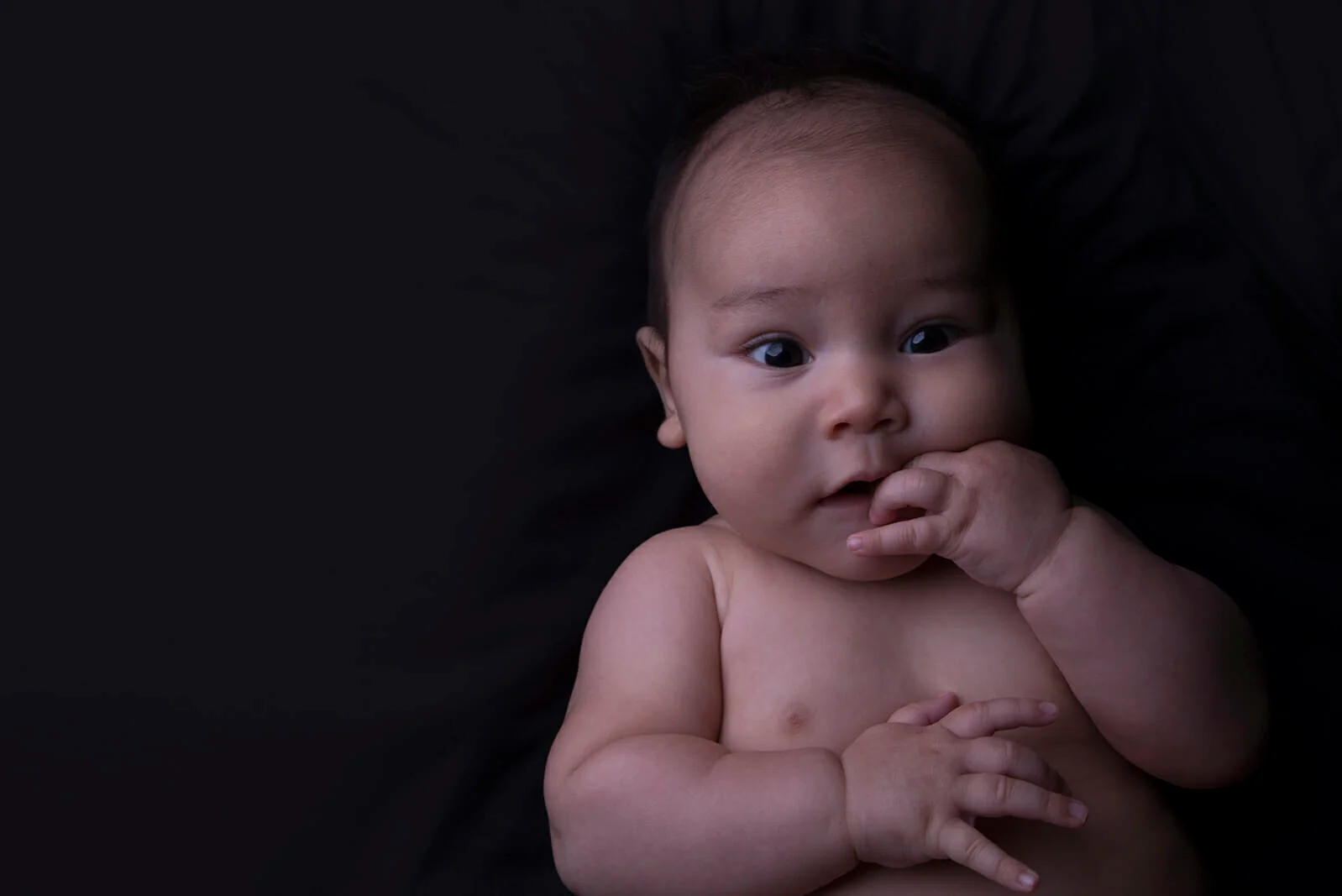 A baby with dark hair lies on a black background, looking up with wide eyes. Perfect for family photography Calgary, the baby is shirtless, with one hand near their mouth and the other resting on their chest. - Jodi O Photography
