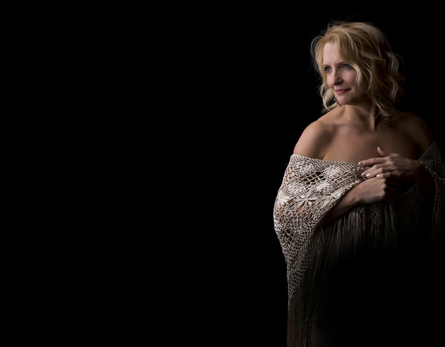 A woman with blonde hair stands against a black background, draped in a fringed, knitted shawl that exposes her shoulders. Captured by a boudoir photographer Calgary, she gazes to the side with a gentle expression, hands delicately holding the shawl. - Jodi O Photography