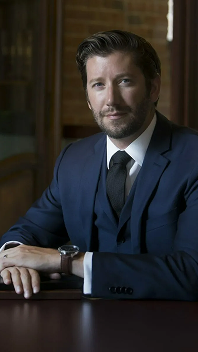 A man with short brown hair and a beard, dressed in a navy blue suit and black tie, sits at a desk with his hands resting on the surface, looking at the camera with a slight smile, captured by a Calgary photographer. - Jodi O Photography