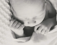 Black and white close-up photo by a Calgary photographer of a newborn baby lying on a soft, textured blanket, eyes closed and hands curled gently near its face. - Jodi O Photography