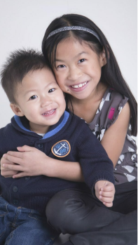 A young girl with long dark hair hugs a smiling toddler boy, both sitting close together and smiling at the camera against a light background, perfectly captured by a Calgary photographer. - Jodi O Photography