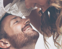 A close-up by a Calgary photographer of a smiling man with a beard and a woman leaning toward him, both appearing happy and affectionate, dressed in light-colored clothing. - Jodi O Photography