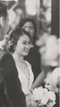 A black-and-white photo by a Calgary photographer captures a smiling bride in a white dress holding a bouquet, standing beside a man in a suit, with another woman slightly behind them at the wedding ceremony. - Jodi O Photography