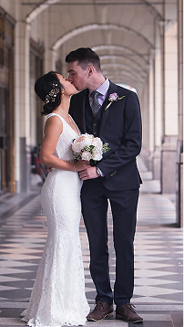 A bride and groom stand close, kissing under a covered walkway with arches. Captured by a Calgary photographer, the bride wears a white lace dress with bouquet; the groom is in a dark suit with boutonnière. - Jodi O Photography