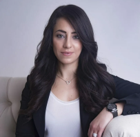 A woman with long dark hair sits on a light-colored couch, wearing a black blazer, white top, and a watch, looking confidently at the camera—captured brilliantly by a Calgary photographer. - Jodi O Photography