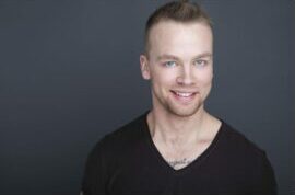 A smiling man with short blonde hair and blue eyes, wearing a black V-neck shirt, poses in front of a dark gray background—perfect for professional actor headshots Calgary style. - Jodi O Photography
